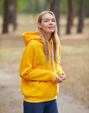 Foto de Atractiva mujer rubia de ojos azules caminar por el parque de la ciudad. Chica llevar sudadera con capucha amarilla, bolsa de color rosa y se ven felices y sonríe. Retrato de una joven alegre disfrutando en el parque de otoño. Relájese en la naturaleza. - Imagen libre de derechos