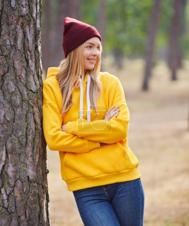 Foto de Rubia de ojos azules con capucha amarilla y sombrero borgoña en un bosque de pinos. Retrato de una joven alegre disfrutando en el parque de otoño. Relájese en la naturaleza. - Imagen libre de derechos