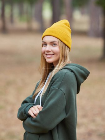 Foto de Rubia de ojos azules con capucha verde y sombrero amarillo en un bosque de pinos. Retrato de una joven alegre disfrutando en el parque de otoño. Relájese en la naturaleza. - Imagen libre de derechos