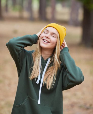 Foto de Rubia de ojos azules con capucha verde y sombrero amarillo en un bosque de pinos. Retrato de una joven alegre disfrutando en el parque de otoño. Relájese en la naturaleza. - Imagen libre de derechos
