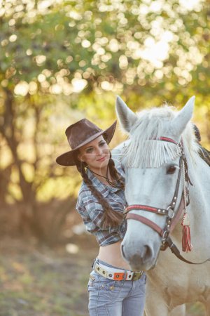 Photo for Happy beautiful young woman cowgirl in hat standing and hugging her horse - Royalty Free Image