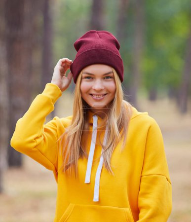 Foto de Atractiva mujer rubia de ojos azules caminar por el parque. Chica llevar sudadera con capucha amarilla y sombrero de color burdeos, se ven felices y sonríe. Retrato de una joven alegre disfrutando en el parque de otoño. - Imagen libre de derechos