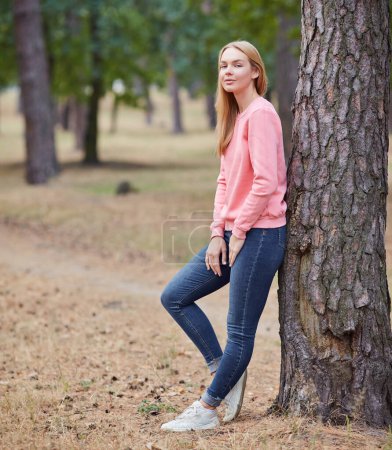 Téléchargez les photos : Blond aux yeux bleus dans un sweat à capuche rose se promène dans une forêt de pins. Portrait d'une jeune femme joyeuse jouissant dans le parc d'automne. Détendez-vous dans la nature. - en image libre de droit