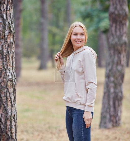Photo for Blue-eyed blonde in a beige hoodie walks in a pine forest. Portrait of a joyful young woman enjoying in autumn park. Relax in nature. - Royalty Free Image