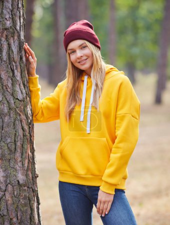 Foto de Atractiva mujer rubia de ojos azules caminar por el parque. Chica llevar sudadera con capucha amarilla y sombrero de color burdeos, se ven felices y sonríe. Retrato de una joven alegre disfrutando en el parque de otoño. - Imagen libre de derechos