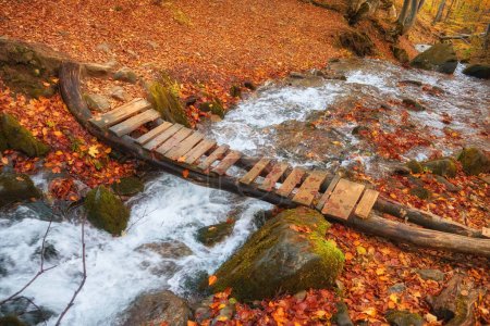 Foto de Serenidad de la escena del bosque de otoño. Un pequeño arroyo serrano serpentea con gracia a través del follaje vibrante, adornado con un encantador puente de madera. - Imagen libre de derechos