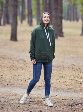 Foto de Atractiva mujer rubia de ojos azules caminar por el parque de la ciudad. Chica usar sudadera con capucha verde, mirada feliz y sonrisas. Retrato de una joven alegre disfrutando en el parque de otoño. - Imagen libre de derechos