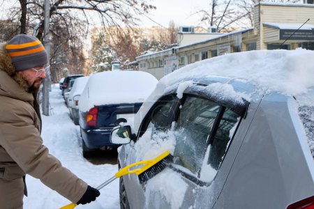 Téléchargez les photos : Un homme nettoie la neige d'une voiture à l'aide d'une brosse par une journée froide d'hiver, mettant en valeur la tâche routinière du déneigement - en image libre de droit