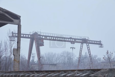 Foto de Una grúa pesada se encuentra en una instalación industrial al aire libre, envuelta en la niebla de la madrugada. - Imagen libre de derechos