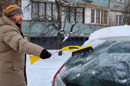 Téléchargez les photos : Un homme nettoie la neige d'une voiture à l'aide d'une brosse par une journée froide d'hiver, mettant en valeur la tâche routinière du déneigement - en image libre de droit