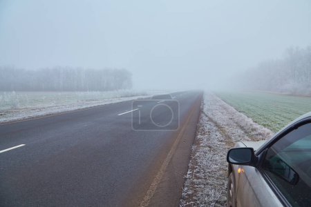 Téléchargez les photos : Une voiture roule le long d'une route couverte de brouillard, recouverte d'une couche de gel, créant une atmosphère de mystère hivernal et de froid. - en image libre de droit
