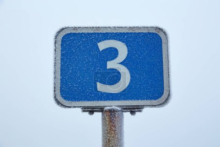 Foto de Una señal de tráfico cubierta de heladas se encuentra en el borde de la carretera, rodeada por un paisaje nublado de invierno.. - Imagen libre de derechos
