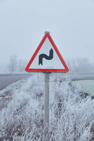 Téléchargez les photos : Un panneau routier recouvert de givre se dresse sur le bord de la route, entouré d'un paysage hivernal brumeux. - en image libre de droit