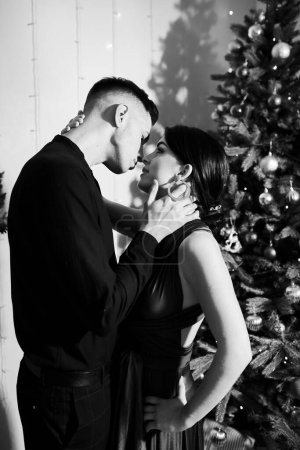 Foto de Una joven pareja enamorada se besa en Nochebuena junto al árbol de Navidad con regalos en casa en diciembre. Foto en blanco y negro - Imagen libre de derechos