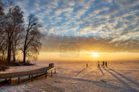 Foto de Una pintoresca noche de invierno en el lago congelado. A medida que el sol se pone, el cielo se convierte en un lienzo de colores ardientes, proyectando un tono cálido sobre el paisaje helado. La gente disfruta de un paseo pausado por la superficie congelada - Imagen libre de derechos