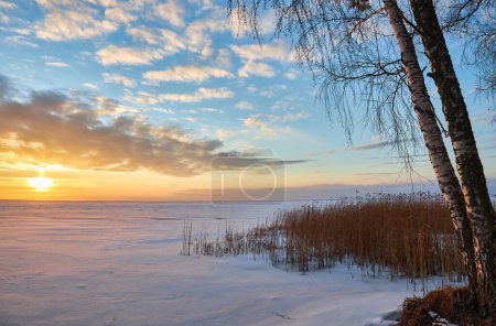 Foto de Una cautivadora escena de invierno con un lago congelado bajo los tonos encantadores del sol poniente. El cielo arde con colores vibrantes, proyectando un resplandor sereno sobre el paisaje helado - Imagen libre de derechos