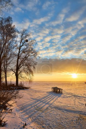 Foto de Una cautivadora escena de invierno con un lago congelado bajo los tonos encantadores del sol poniente. El cielo arde con colores vibrantes, proyectando un resplandor sereno sobre el paisaje helado - Imagen libre de derechos