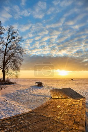 Foto de Una impresionante escena de invierno con un tranquilo lago congelado. A medida que el sol se pone, el cielo se transforma en una obra maestra de colores vibrantes, proyectando un cálido resplandor naranja sobre el paisaje helado y un encantador muelle de madera.. - Imagen libre de derechos