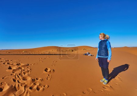 Téléchargez les photos : Un voyageur solitaire au Sahara, captivé par la vaste étendue de dunes dorées sous le soleil brillant - en image libre de droit