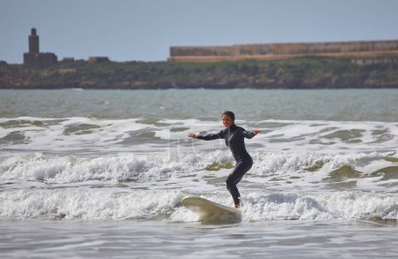 Téléchargez les photos : Une jeune surfeuse chevauche gracieusement les vagues sur sa planche le long des rives d'Essaouira, au Maroc, capturant le frisson et la beauté de la culture du surf contre le littoral pittoresque. - en image libre de droit