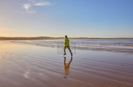 Photo for A young man in a green jacket enjoys a run along the Essaouira coastline in Morocco, where the ocean breeze invigorates the seaside atmosphere - Royalty Free Image