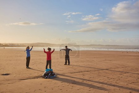 Téléchargez les photos : Les jeunes adultes dynamisent leur journée avec une séance d'entraînement en groupe au bord de l'océan à Essaouira. L'ambiance côtière d'Essaouira au Maroc encadre leur routine matinale revigorante - en image libre de droit