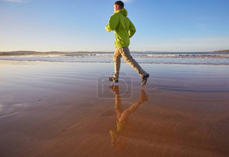Téléchargez les photos : Un jeune homme vêtu d'une veste verte profite d'une course le long du littoral d'Essaouira au Maroc, où la brise de l'océan vivifie l'atmosphère balnéaire - en image libre de droit