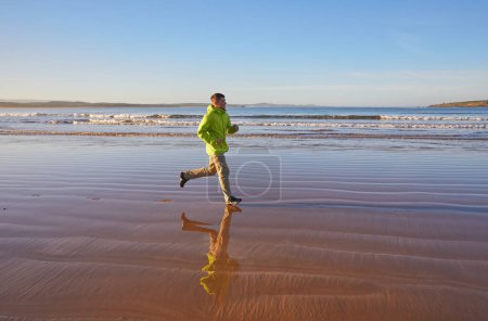 Téléchargez les photos : Un jeune homme vêtu d'une veste verte profite d'une course le long du littoral d'Essaouira au Maroc, où la brise de l'océan vivifie l'atmosphère balnéaire - en image libre de droit