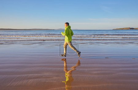 Foto de Un joven con una chaqueta verde disfruta de una carrera a lo largo de la costa de Essaouira en Marruecos, donde la brisa del océano vigoriza la atmósfera costera - Imagen libre de derechos