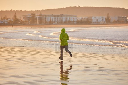 Foto de Un joven con una chaqueta verde disfruta de una carrera costera a lo largo del océano de Essaouira, las olas que proporcionan una banda sonora refrescante a su vigorizante trote. - Imagen libre de derechos