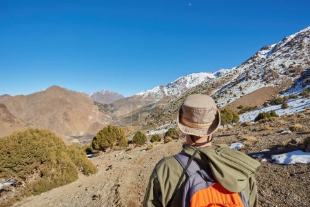 Foto de Un turista se para en el paso de las montañas del Atlas, contemplando picos nevados, capturando la majestuosidad de esta serena escapada de montaña. - Imagen libre de derechos