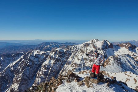Foto de Un turista triunfante en la cima del Monte Toubkal, Marruecos, celebrando el ascenso con las manos levantadas y un hacha de hielo. - Imagen libre de derechos