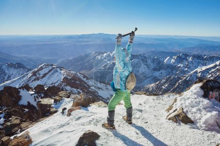 Téléchargez les photos : Un touriste triomphant au sommet du mont Toubkal, au Maroc, célébrant l'ascension avec les mains levées et une hache de glace. - en image libre de droit