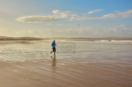 Téléchargez les photos : Une jeune femme profitant d'un jogging côtier le long du front de mer d'Essaouira, avec les vagues de l'Atlantique offrant une toile de fond sereine à sa course revigorante - en image libre de droit