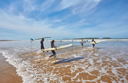 Foto de Una vibrante banda de jóvenes surfistas se precipita en el océano, tablas de remolque, abrazando la emoción de encontrarse con las olas de frente. Cada paso se hace eco del entusiasmo de una aventura surfera que se desarrolla - Imagen libre de derechos