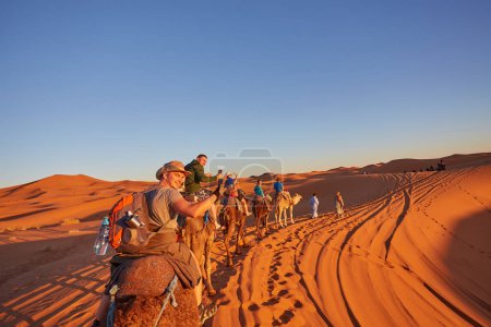 Photo for A group of cheerful tourists embarks on a camel safari, exploring the enchanting Sahara Desert in Morocco - Royalty Free Image