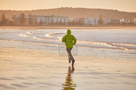 Photo for Energetic young man in a green jacket jogging along the ocean in Essaouira, Morocco. Enjoying the scenic coastline, embracing an active lifestyle - Royalty Free Image