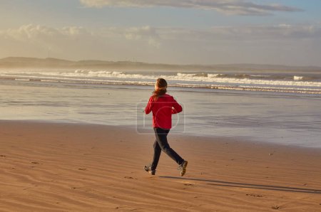 Foto de Una joven disfrutando de una refrescante carrera a lo largo de la orilla del océano en Essaouira, Marruecos, con olas rompiendo suavemente contra el telón de fondo costero - Imagen libre de derechos