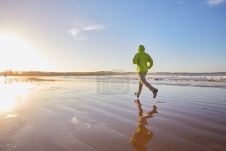 Foto de Un joven con una chaqueta verde disfruta de una carrera costera a lo largo del océano de Essaouira, las olas que proporcionan una banda sonora refrescante a su vigorizante trote. - Imagen libre de derechos