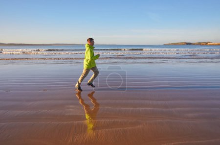 Foto de Joven enérgico con una chaqueta verde corriendo a lo largo del océano en Essaouira, Marruecos. Disfrutando de la costa escénica, abrazando un estilo de vida activo - Imagen libre de derechos