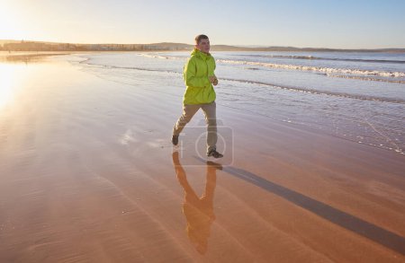 Foto de Joven enérgico con una chaqueta verde corriendo a lo largo del océano en Essaouira, Marruecos. Disfrutando de la costa escénica, abrazando un estilo de vida activo - Imagen libre de derechos