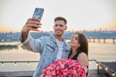 A young couple with a massive bouquet of roses takes a selfie on a rooftop, capturing the cityscape at sunset as the backdrop.