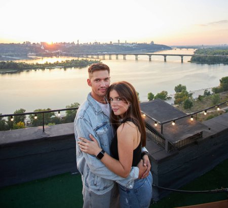 A young couple kisses and embraces during a romantic rooftop date, with a cityscape backdrop at sunset.