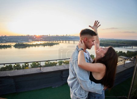 Téléchargez les photos : Un couple partage un moment tendre sur un toit, leurs silhouettes gravées sur le ciel du soir. L'éclat de la ville renforce l'atmosphère romantique. - en image libre de droit
