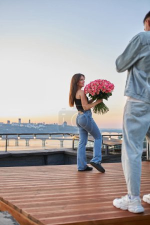 Photo for A young man surprises his beloved with a vast bouquet of roses as they capture their joy in a selfie against the urban sunset on a rooftop.. - Royalty Free Image