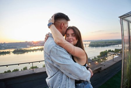 Téléchargez les photos : Un couple partage un moment tendre sur un toit, leurs silhouettes gravées sur le ciel du soir. L'éclat de la ville renforce l'atmosphère romantique. - en image libre de droit