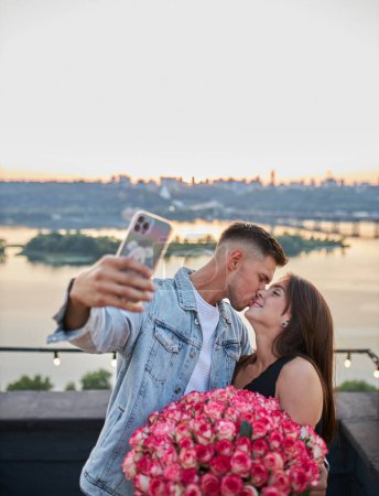 A young man surprises his beloved with a vast bouquet of roses as they capture their joy in a selfie against the urban sunset on a rooftop