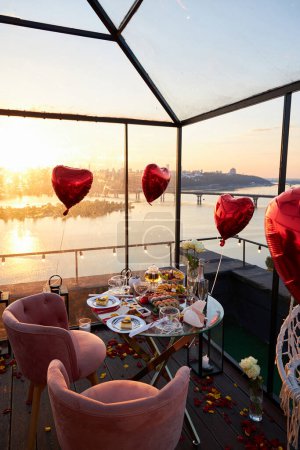 Téléchargez les photos : Dîner romantique sur un toit avec ballons rouges, vue sur la ville au coucher du soleil. - en image libre de droit