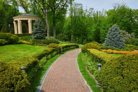Photo for Trimmed green bushes form a labyrinth along tiled pathways in the park, creating a serene and maze-like atmosphere. A perfect summer day unfolds in this beautifully landscaped outdoor space. - Royalty Free Image