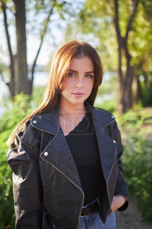A young brunette, adorned in a leather jacket, poses against the backdrop of nature's beauty.